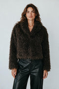 Model wearing a Plaza d'Amanté dark brown shaggy faux fur cropped jacket with a notched collar over black trousers.