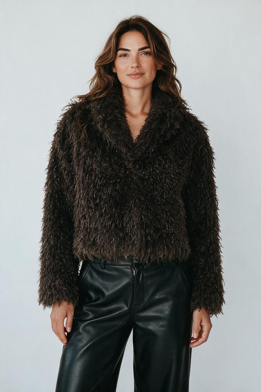 Model wearing a Plaza d'Amanté dark brown shaggy faux fur cropped jacket with a notched collar over black trousers.