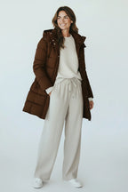 A woman wearing a Plaza d'Amanté chocolate brown longline insulated hooded puffer jacket over a cream sweater and pants.