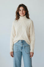 Model wearing a Plaza d'Amanté cream turtleneck sweater with diamond cable knit texture and ribbed cuffs
