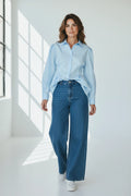 Plaza d'Amanté light blue cotton blend button-down shirt with puff shoulders worn with wide-leg jeans by a window.