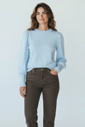 Plaza d'Amanté light blue crew neck sweater with puff sleeves and faux-pearl button cuffs worn by a smiling woman.
