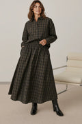 A woman wearing a Plaza d'Amanté plaid button-down shirt and matching midi skirt in earthy tones with black boots.