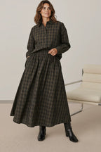 A woman wearing a Plaza d'Amanté plaid button-down shirt and matching midi skirt in earthy tones with black boots.