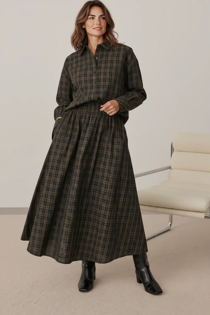 A woman wearing a Plaza d'Amanté plaid button-down shirt and matching midi skirt in earthy tones with black boots.
