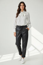 Plaza d'Amanté model wearing a white polka dot tie neck blouse and black faux leather joggers with white sneakers.
