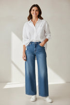 Model wearing a Plaza d'Amanté white button-down shirt and medium-wash high-rise wide-leg denim jeans with sneakers.